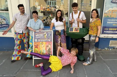 A group of people pose with a poster for the Wallace & Gromit Grand Appeal. They are all smiling and wearing colourful trousers as part of the Wrong Trousers Day in Bristol
