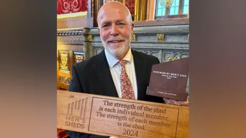 Simon is bald and is wearing a black suit jacket, white shirt and pink and white floral tie. He has a white moustache and beard, and is holding a large wooden plaque that says "the strength of the shed is each individual member. The strength of each member is the shed." He is also holding a small brown trophy.