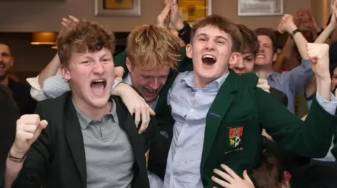 A group of young people celebrating in a golf club supporting Rory McIlroy. Two of them are wearing green blazers and collared shirts. Another is wearing a green polo shirt. They're cheering with their arms in the air