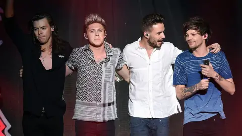 Getty Images Harry Styles, Niall Horan, Liam Payne and Louis Tomlinson standing in a row. Harry is wearing a black suit and has long brown hair. Niall has blonde short hair and a black patterned shirt. Liam is wearing jeans and a white shirt. Louis has brown short hair and is wearing a blue t shirt, holding a microphone