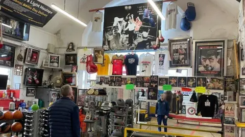 The image is a colourful collection of boxing equipment and memorabilia. Windows on the left and centre walls are just visible with t-shits and boxing photographs and a large pair of blue boxing gloves hanging. Glyn Rhodes stands slightly left of the picture wearing dark blue gym clothing and facing his reflection in the distance, around him to the left side of the room are an array of weights including hand and kettle weights.