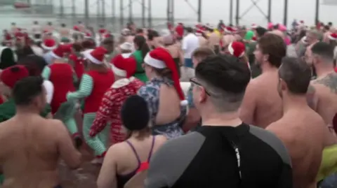 BBC People wearing red Santa hats and swimming costumes heading into the sea for their Christmas Day swim.