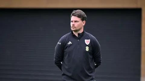 Football Association of Wales Drew Sherman standing with hands behind his back and looking to the side - he's wearing black Welsh training kit