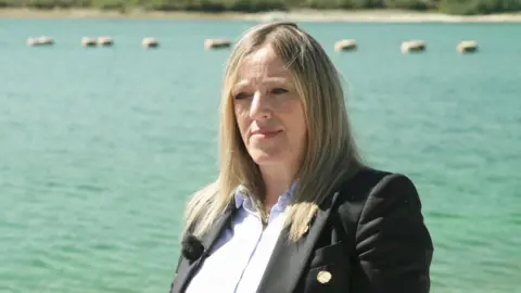 Susan Davy pictured in front of a large reservoir on a sunny day. She is wearing a light blue blouse with a dark coloured blazer. She has straight long dark blonde hair. 