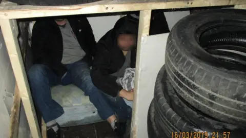 Home Office Two men are sitting inside a confined compartment constructed with wooden panels in the back of a van. The men are partially obscured, wearing dark clothing, and one has a blurred face. The compartment is surrounded by stacked car tyres, and a mattress is visible under the men.