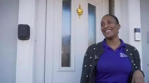 WMCA Sasha Beckford stands outside her 1920s home with the front door closed behind her. It has glass panes and a door knocker. She is smiling, looking happy, and is wearing a purple top with a black cardigan.