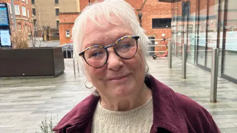 Danielle Stone with short white hair and brown-framed glasses. She is smiling at the camera, and wearing a white sweater and red coat. There is a glass-fronted office building to the right, a grey-slabbed pavement and a wooden planter to the left. There are brick buildings behind