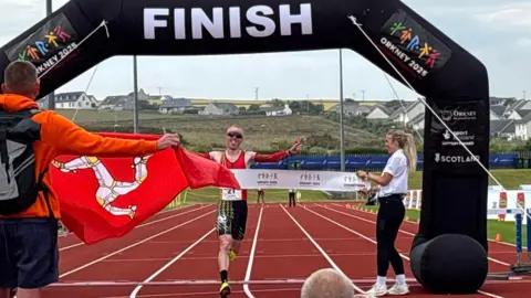 Corrin Leeming raises his arms as he crosses the finish line on an athletics track under an inflatable black arch. He wears a red and black lycra running top and shorts, sunglasses, and yellow trainers. A woman with blonde hair holds up a finishing line ribbon that he runs towards. A spectator holds up a red Manx flag nearby.