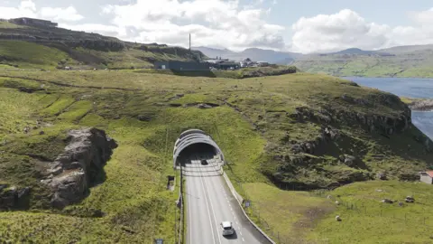 Drone shot of a white car preparing to enter a tunnel in the Faroes. The tunnel has been constructed under green fields and some sheep are also visible in the image.