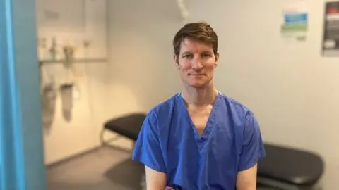 Tim Lawrence is wearing blue medical scrubs and sat in a clinic room