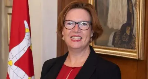 IOM GOV Jane Poole-Wilson standing nest to a Manx flag