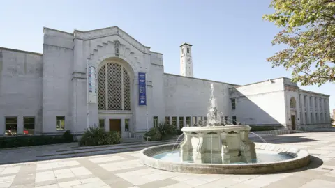 The front facade of the gallery, which is a pale stone building with a cathedral like shape at the front, complete with a large glass section. Outside of the front door is a fountain.