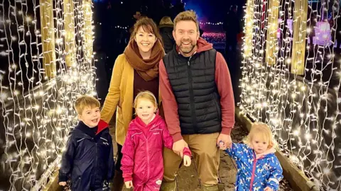 Katie and the Tom family stand under an archway of fairy lights. Katie and Tom have three children, a boy and two girls.