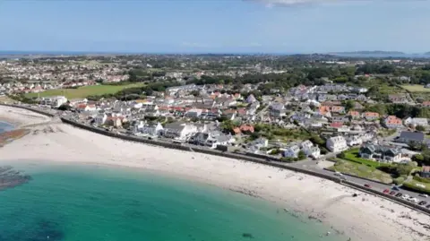 A drone image of Guernsey with the sea on the bottom with sand and houses further back. 