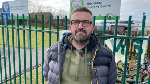 Man stands by a green fence outside a school. He has brown hair and a beard, which is greying. Signs in the background read 'Queensway Primary School & Nursery' and 'Aireborough Children's Centre'.