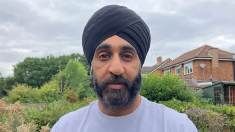 Mr Singh has a black turban and has a dark beard. He is wearing a white T-shirt.