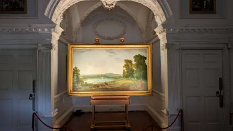 Betsy Ogilvie A painting of a rural setting which includes a lake and some trees. It is illuminated between white pillars and white walls.