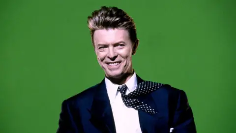 David Bowie is pictured in a navy blue suit, with a white shift and navy and white tie, in front of a bright green background. He's smiling, and the tie is flipped back over one shoulder