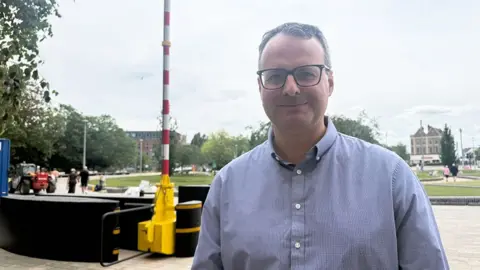 Sarah-May Buccieri/BBC A man with short dark hair and wearing glasses and a light blue shirt. He is standing in front of one of the security barriers in front of the park.