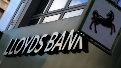 An exterior shot of a branch of Lloyds bank showing a sign with the bank's name and another smaller sign with it's logo of a black horse.