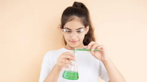 Young indian girl student doing science experiment mixing colorful chemical in a flask isolated over beige background. Education concept.