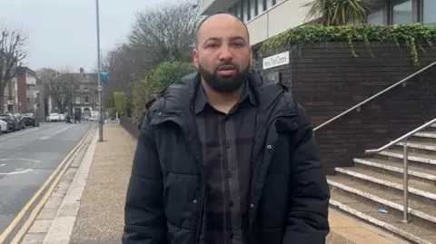 BBC News Jay Lindo looks outside Hove Trial Centre. He wears a black puffer jacket and a dark checked shirt