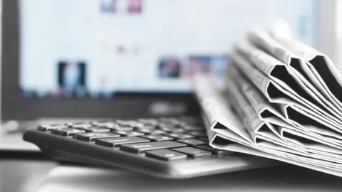 Getty Images A stack of folded newspapers sits on a keyboard in front of a monitor showing a blurred news website