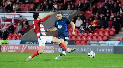 Chris Vaughan/Getty Images Ben House, wearing a dark blue football kit, takes a shot towards goal as a player wearing red and white stripes slides in for a tackle. Red seats, supporters and advertising boards are in the background.