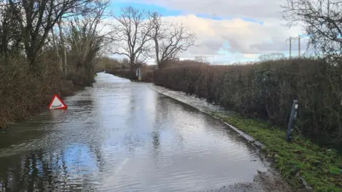 Weather watcher Dell Boy Flooded road at Sturminster Marshall in Dorset. A flood street sign is almost submerged in the water.