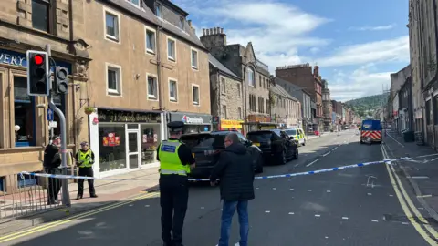 A police cordon on the street in Perth. A police officer in a hi-viz vest is speaking to a man in a dark jacket and jeans. They are standing in front of a strip of police tape.
