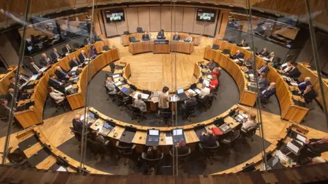 Senedd members seated at their desks in the parliament's circular chamber.
