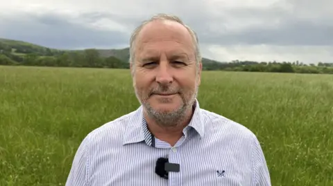Mike Rigby is standing in a green field wearing a blue and white pin-striped shirt. He has short grey hair and a short beard.