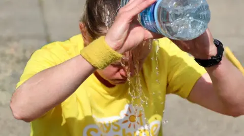 Naomi Weir A close up of Lucy Bird sitting on the ground wearing a bright yellow T-shirt. Her head is down and she is pouring water from a large plastic bottle onto her head and face. 
