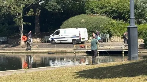 Quewone Bailey-Fleet A white van is parked at the side of a lake with lots of people holding pipes running into the water.