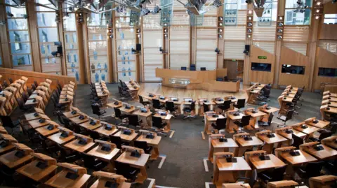 Getty Images The large, modern legislative chamber at the Scottish Parliament - with curved rows of desks and black chairs arranged in a semi-circular layout. Tall windows, wooden beams, and numerous overhead lights create a bright, spacious environment. At the centre stands a raised podium area, likely used for speeches or official proceedings.