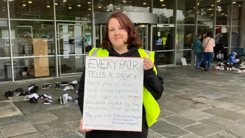 Kirsty Pellow is wearing a black hoodie, black trousers and a green hi-viz vest. She  has shoulder length brown hair. She is holding a whiteboard which says "EVERY PAIR TELLS A STORY." Behind her is County Hall, a building made of glass with a glass revolving door. On the grey concrete are children shoes, behind her to the left and right. There are people to the right of her in the background. 