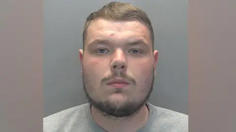 Reis Treby is looking at the camera in a police custody photo. He has short, light brown hair and a beard and moustache. He is wearing a grey top