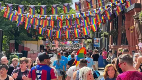 A huge crowd of people walks through a street. Rainbow LGBTQI+ flags hand from the buildings overhead. 