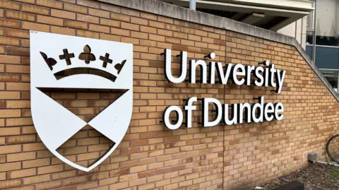A brown brick wall with the words University of Dundee spelled out in silver letters on the wall. Also on the wall, next to the words, is the university's crest. 