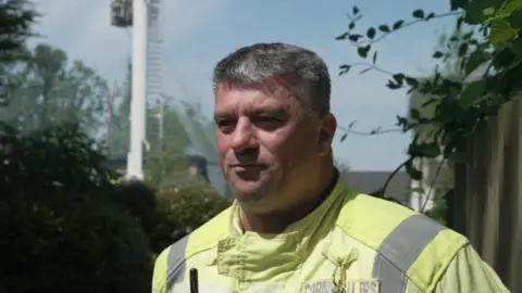 Jason Doney wearing a high viz yellow jacket. He is standing in front of the scene and has short grey hair. He is clean shaven.