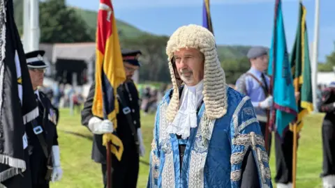 Laurence Skelly in the president's elaborate blue robes and wig walking past a row of standard bearers.
