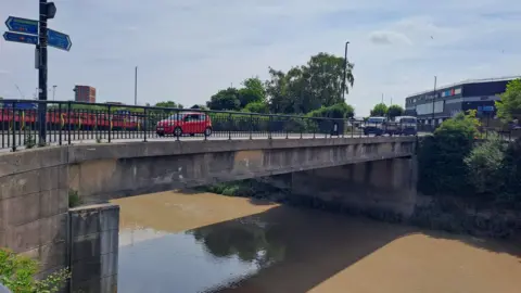 A bridge running over a river with brown water. A red car is driving over the bridge.