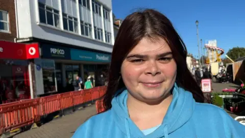 Andrew Turner/BBC Amy Newey smiles directly at the camera. She has long black hair and wears a blue hoodie. It is located near Poundland in Great Yarmouth, where there are orange pedestrian barriers in the street due to groundworks.