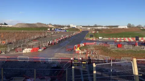 A view of intersecting roads around the site of the HS2 construction around Kenilworth, Warwickshire. A series of barriers can be seen in the foreground and away in the distance, where a large pile of gravel can be seen.