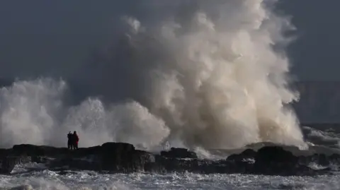 Reuters Large waves smash against rocks along the coast of Porthcawl, Wales, as two people look on