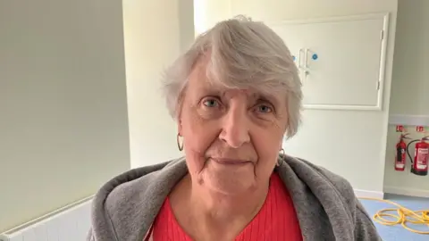 Sam Read/BBC Carol Wilson with medium-length grey hair and large gold earrings. She is wearing a red top and grey coat. There is a white cupboard and fire extinguishers behind her.