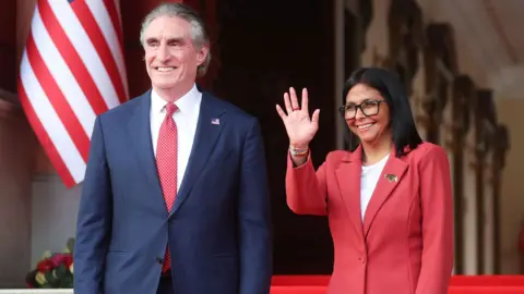 Venezuela's interim president, Delcy Rodriguez and US Interior Secretary Doug Burgum stand side by side smiling and Delcy Rodriguez waves