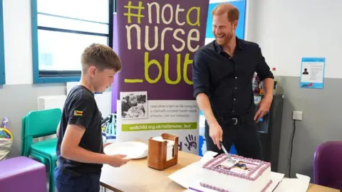 PA Media Prince Harry rests a knife on a cake and laughs, a twelve-year-old boy's eyes are firmly focused on the cake. 