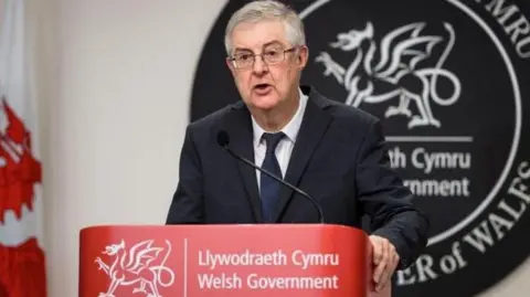 Getty Images Mark Drakeford standing at a Welsh government branded lectern hosting a news conference during the pandemic. He has a large circular grey bilingual sign made of slate behind him saying Welsh government and first minister of Wales and a Welsh flag to his right