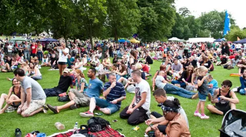 REDFEST A crowd of people, some sitting, others standing, in a lush green grass in a bright day, during a summer festival